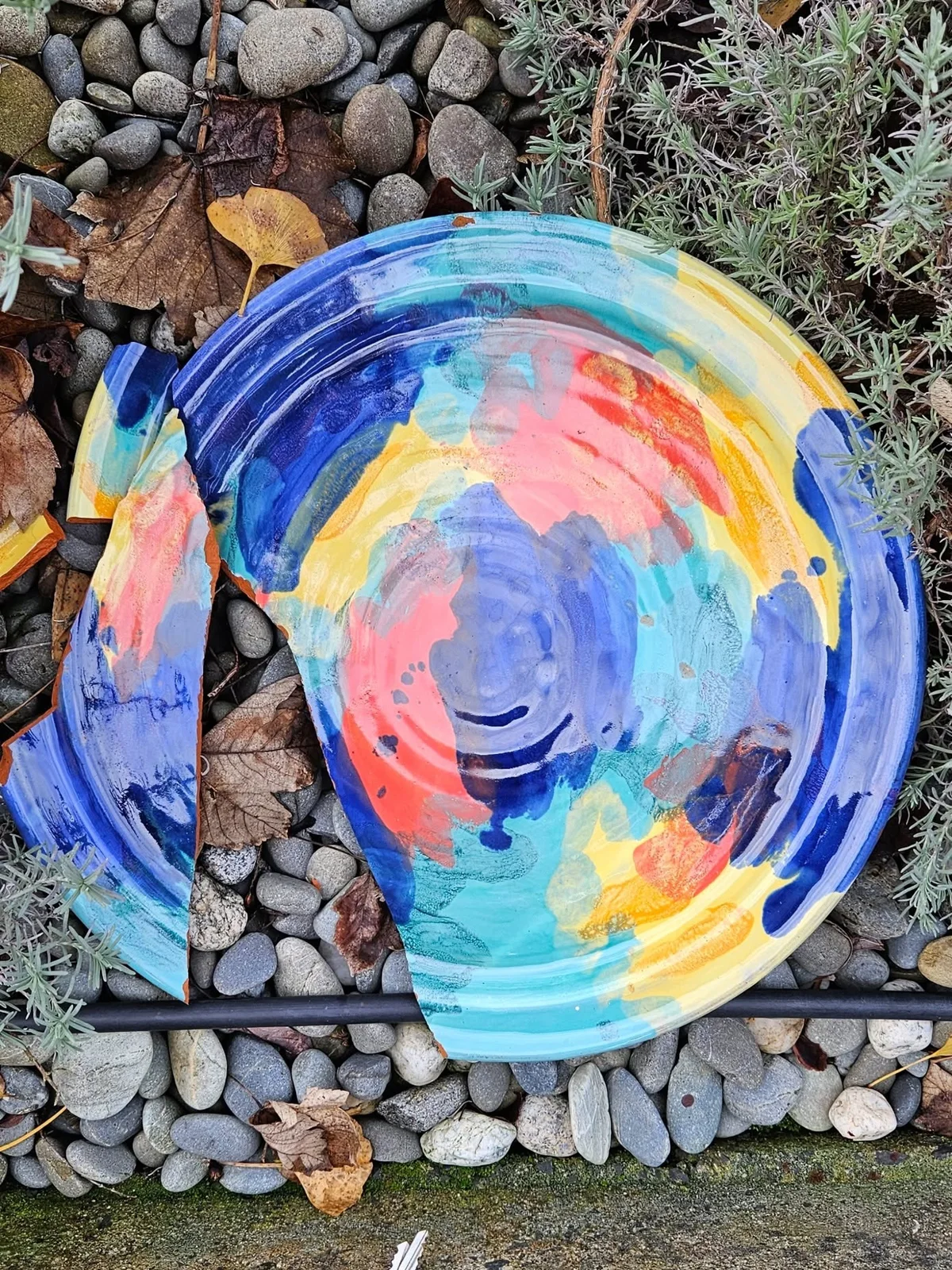 A round dish painted in blue, turquoise, yellow and coral, broken in two on garden stones