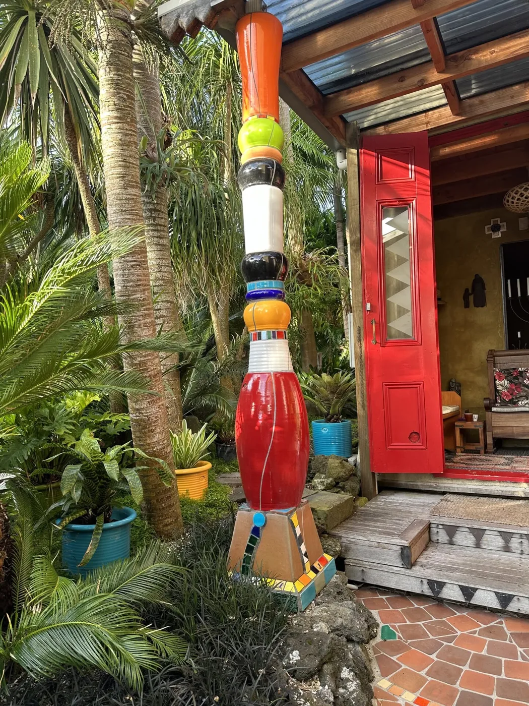 A tall stacked column of glazed Morris & James pieces in orange, lime, black, white and red, in a tropical garden beside a red door