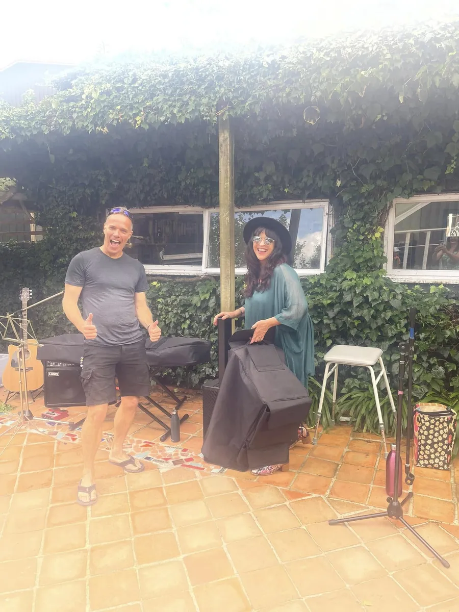 Two people laughing and celebrating beside music gear in the courtyard, under the vine-covered pergola