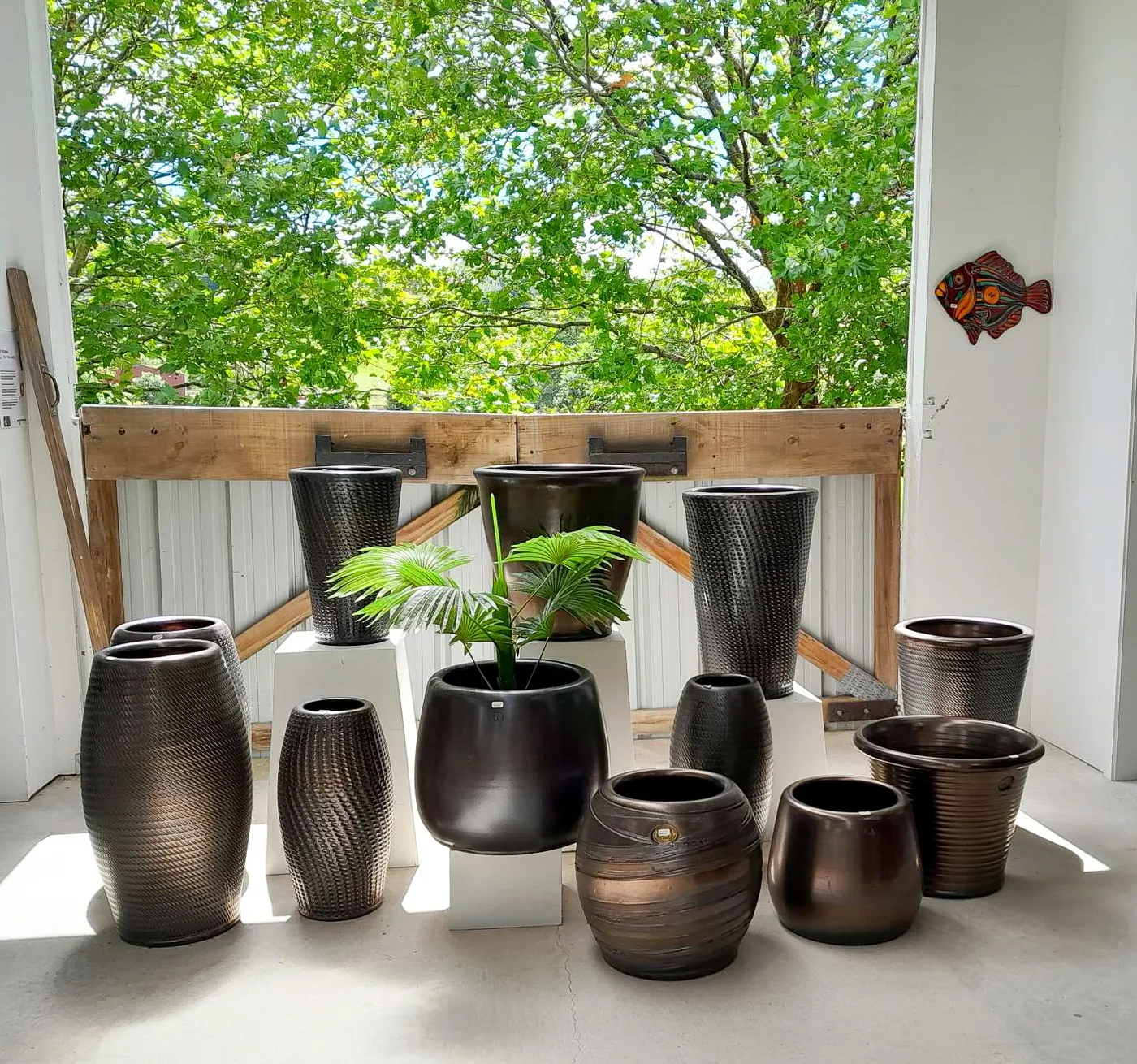 A showroom display of bronze-glazed pots and vases in textured, roulette and plain shapes, in front of an open door to the trees